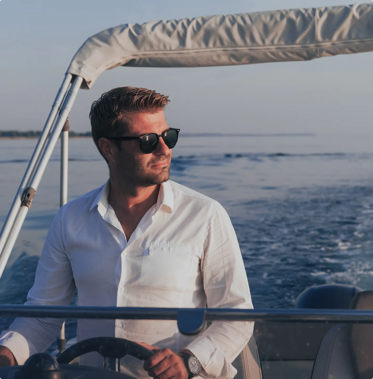 Man in a white shirt and sunglasses steering a boat on calm water during sunset.