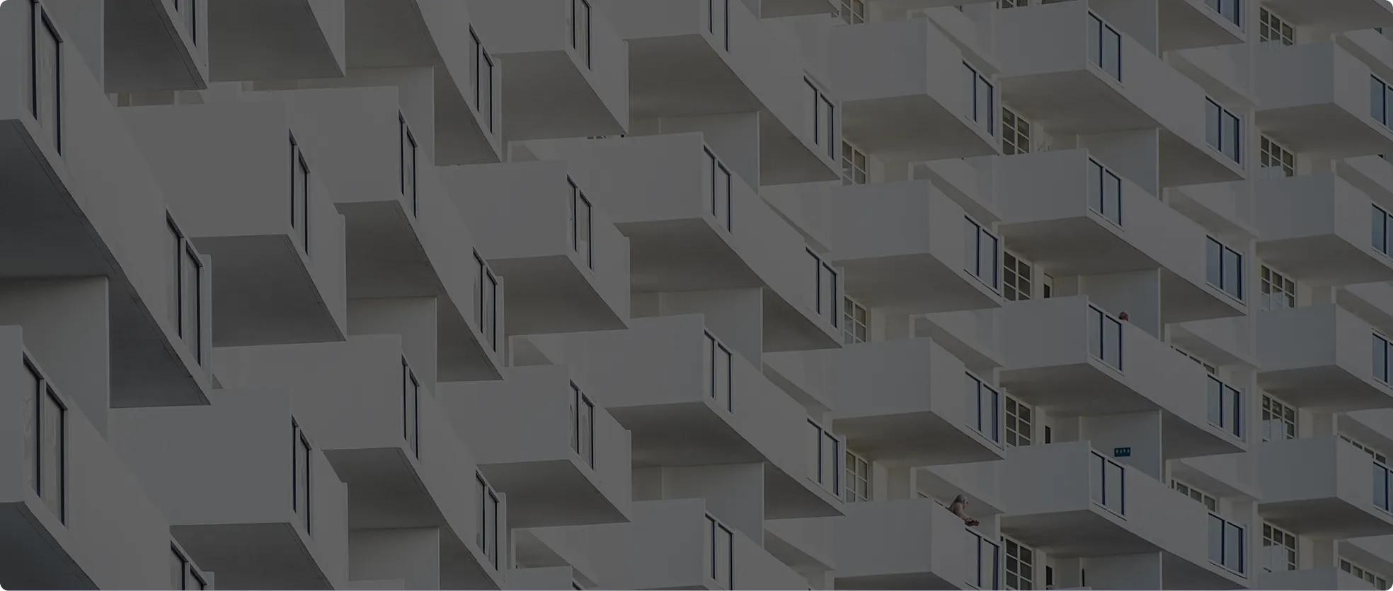 View of a modern building façade featuring rows of white balconies with glass railings arranged in a staggered pattern, seen from an angle.