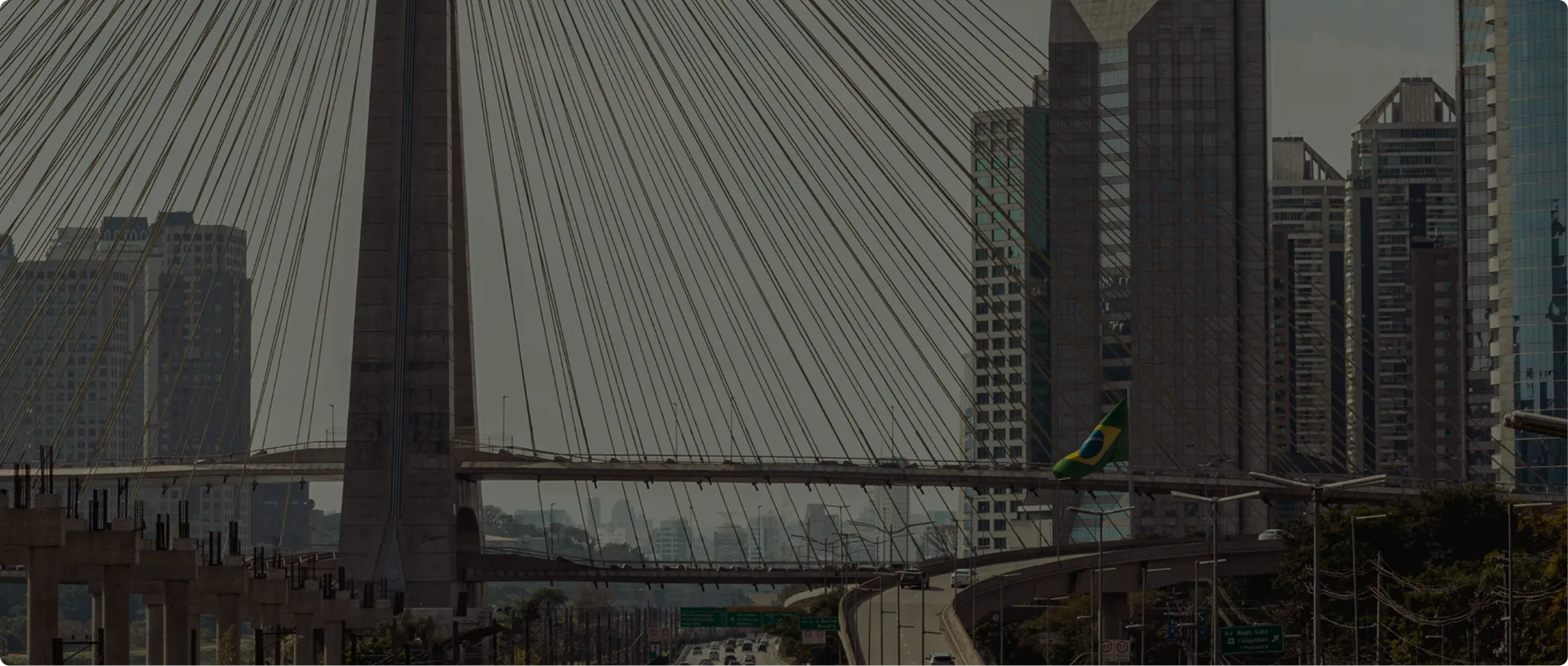 Cable-stayed bridge spanning over a highway with multiple lanes of vehicles, surrounded by tall modern buildings, including one with a Brazilian flag.