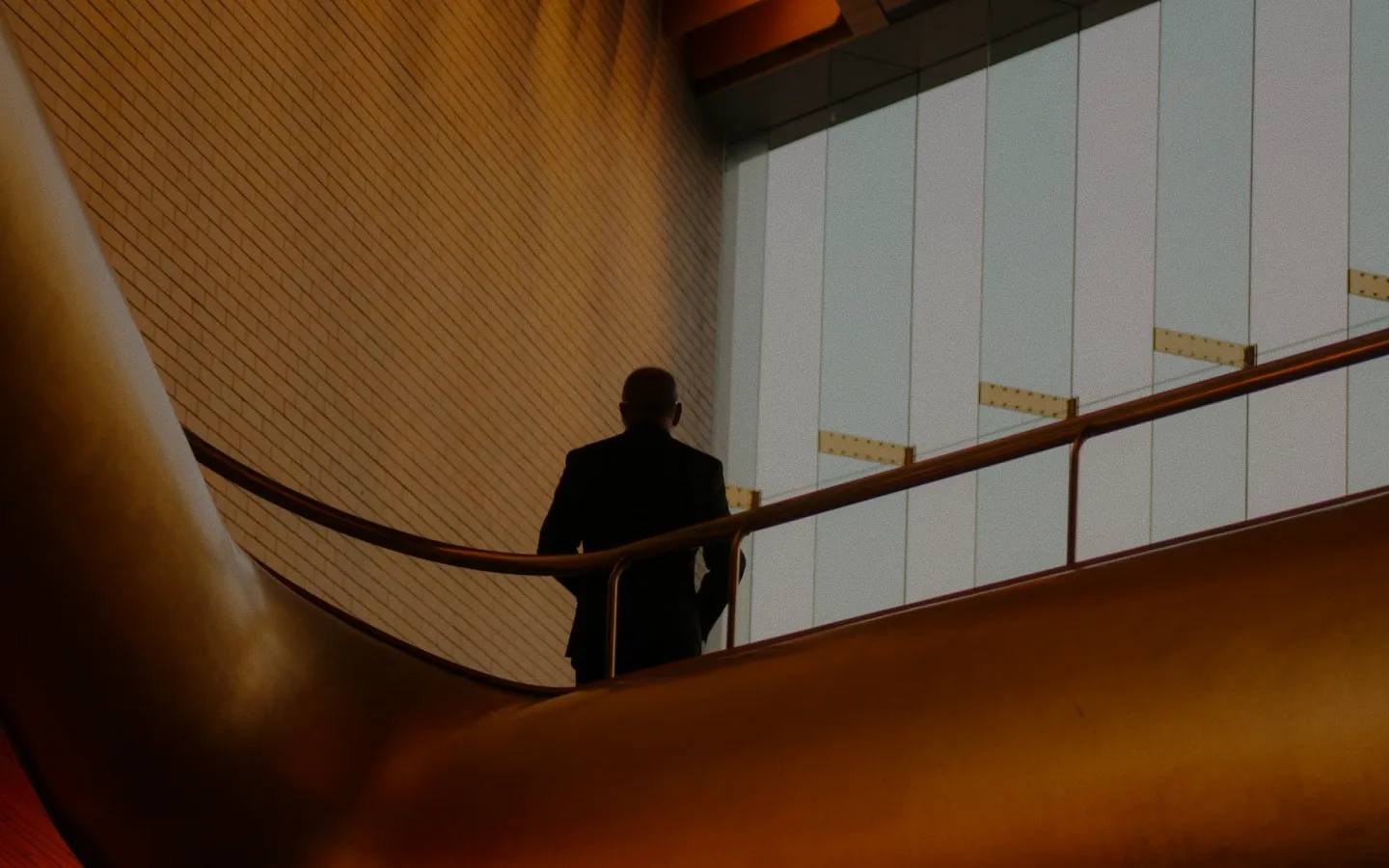 Silhouette of a person in dark clothing standing on an indoor staircase with modern architectural design featuring a warm-toned curved railing and a light-colored tiled wall.