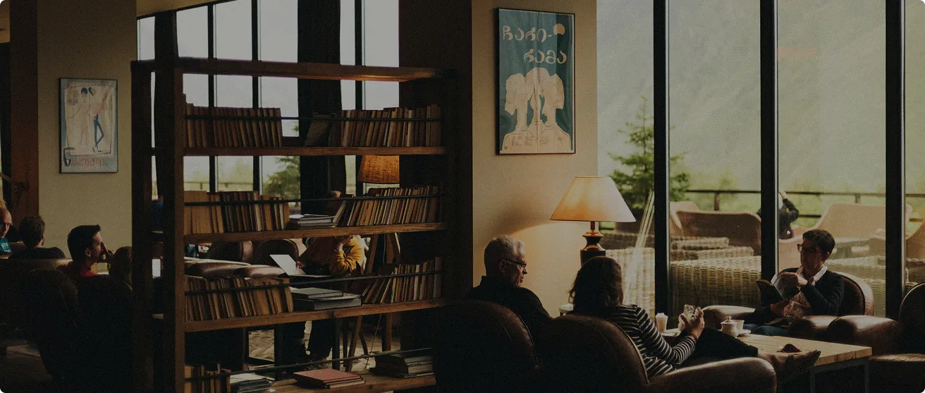 Cozy indoor lounge with large windows showing outdoor seating; people sit on leather chairs and sofas reading or working near wooden bookshelves and a table lamp.