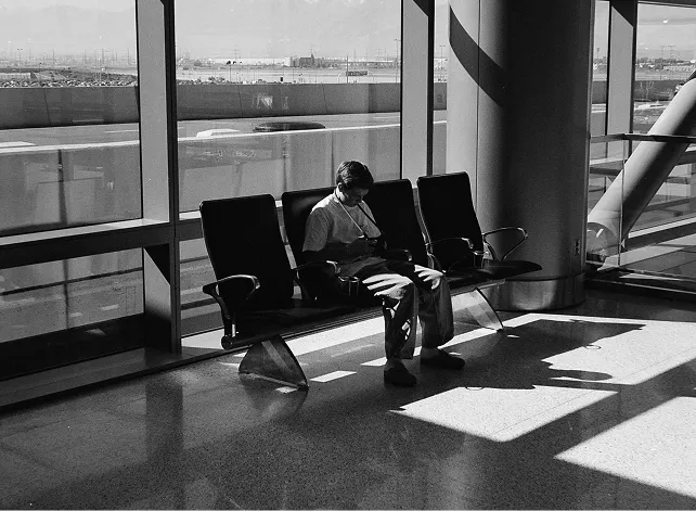 A person sitting alone on airport waiting area seats next to a large window, looking down at a phone or device, with sunlight casting shadows on the floor.