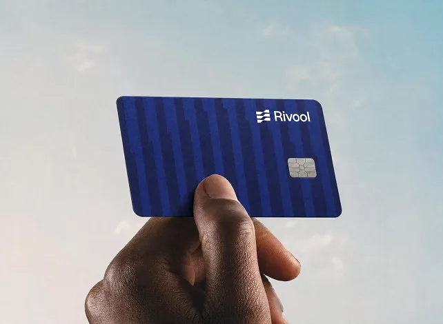 Close-up of a hand holding a blue striped Rivool credit card with a chip against a cloudy sky background.