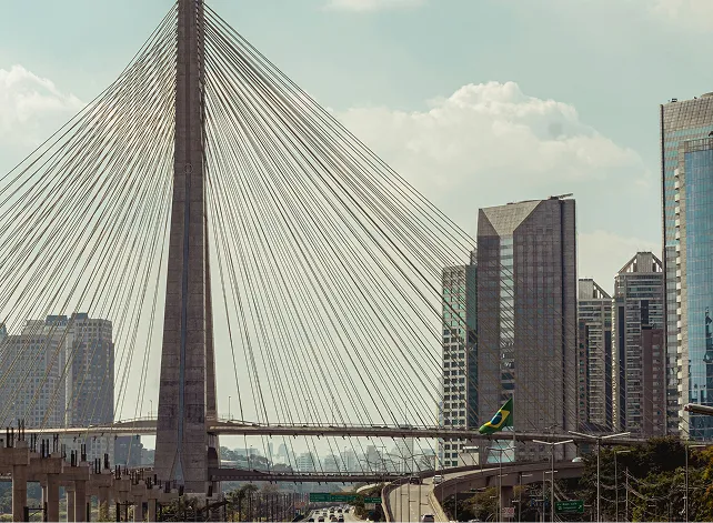 Cable-stayed bridge with a tall central pylon and numerous supporting cables against a cloudy sky, with a cityscape of modern skyscrapers in the background.