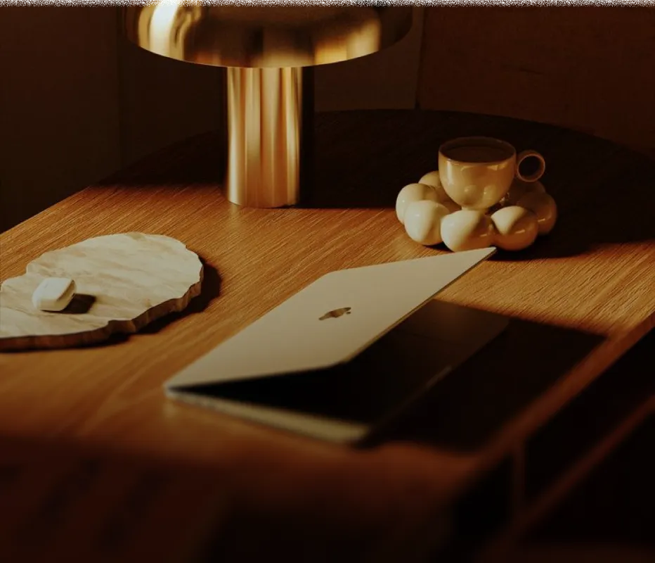 Wooden desk with a partially closed silver laptop, a modern gold lamp, a small textured stone tray with a white object, and a white cup on a unique rounded bubble-style saucer holding a dark beverage.