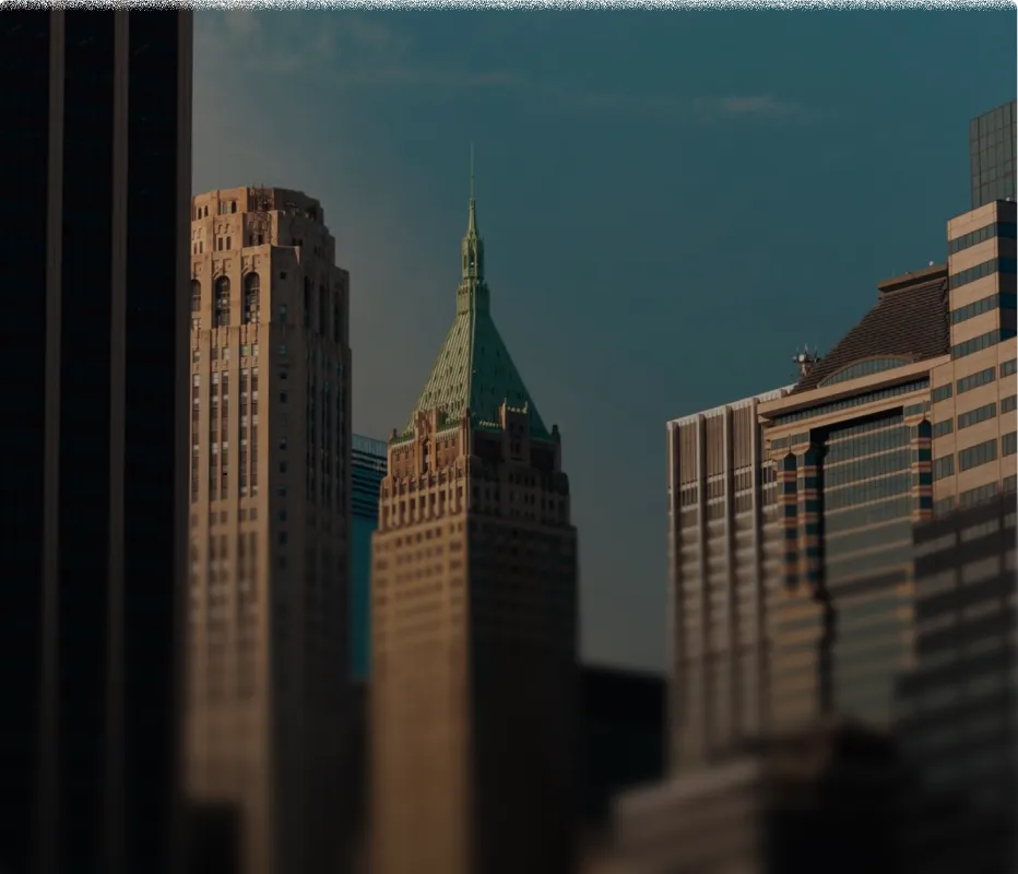 A cityscape featuring multiple tall buildings with one prominent building having a green pointed roof illuminated by sunlight against a dark sky.