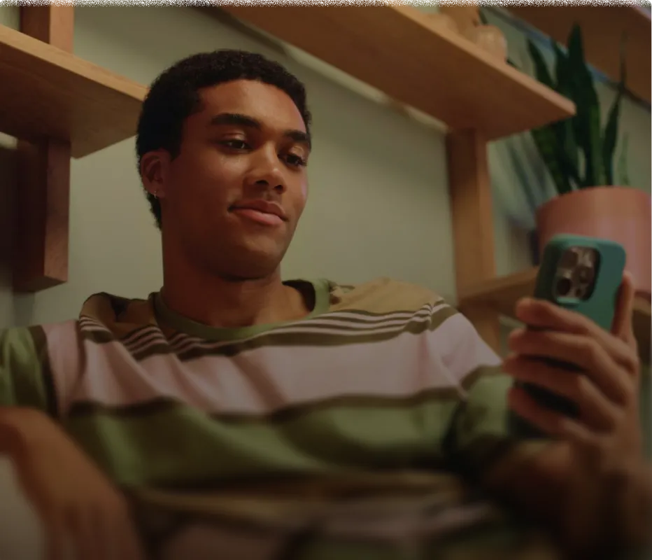 Young man wearing a striped shirt looking at a smartphone with a neutral expression while sitting indoors near wooden shelves and a potted plant.
