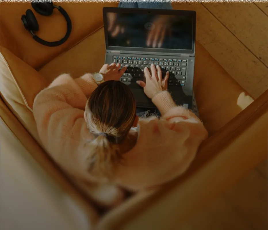 Person with light brown hair tied in a ponytail wearing a fuzzy light pink sweater, typing on a laptop while sitting on a brown couch with black headphones nearby.