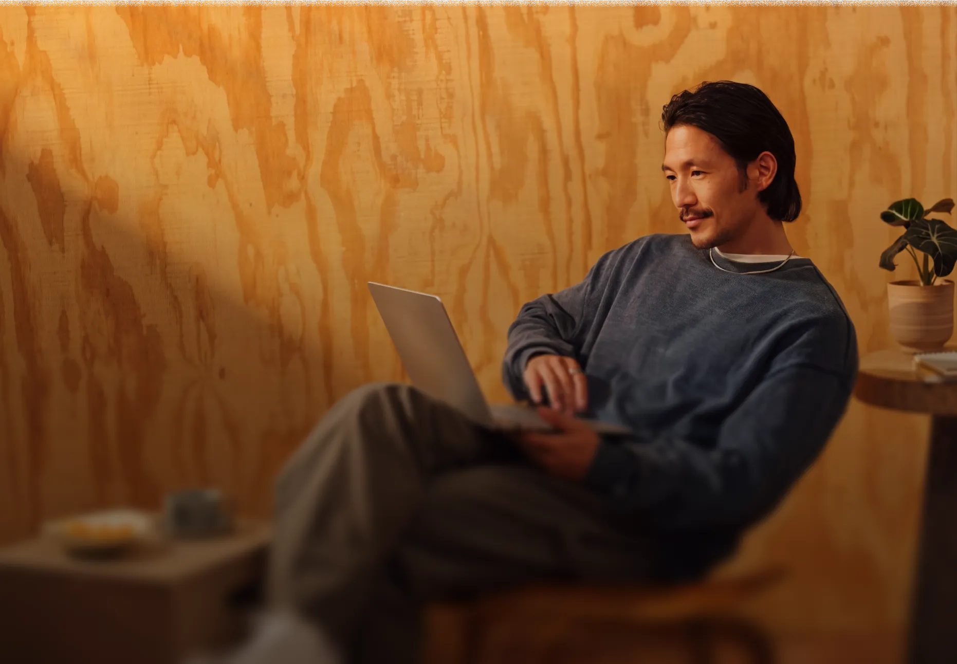 A man in a blue sweater sits on a wooden chair using a laptop in a warm wood-paneled room with a small plant on a round table nearby.