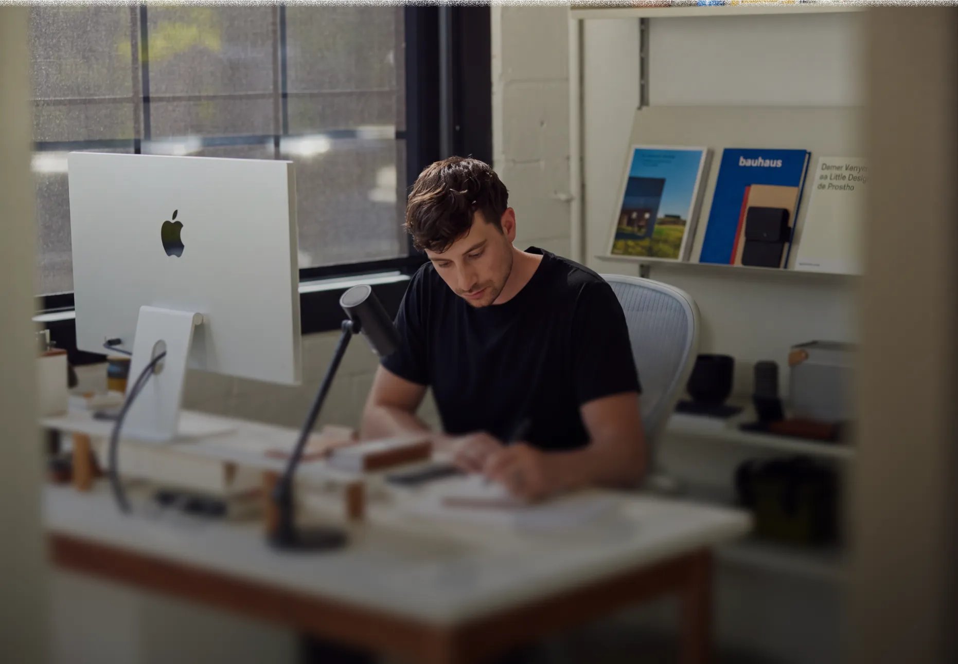 A man in a black shirt sits at a desk in an office, writing on paper with a pen, with a large Apple desktop computer and a microphone on the desk, and shelves with books and objects in the background.