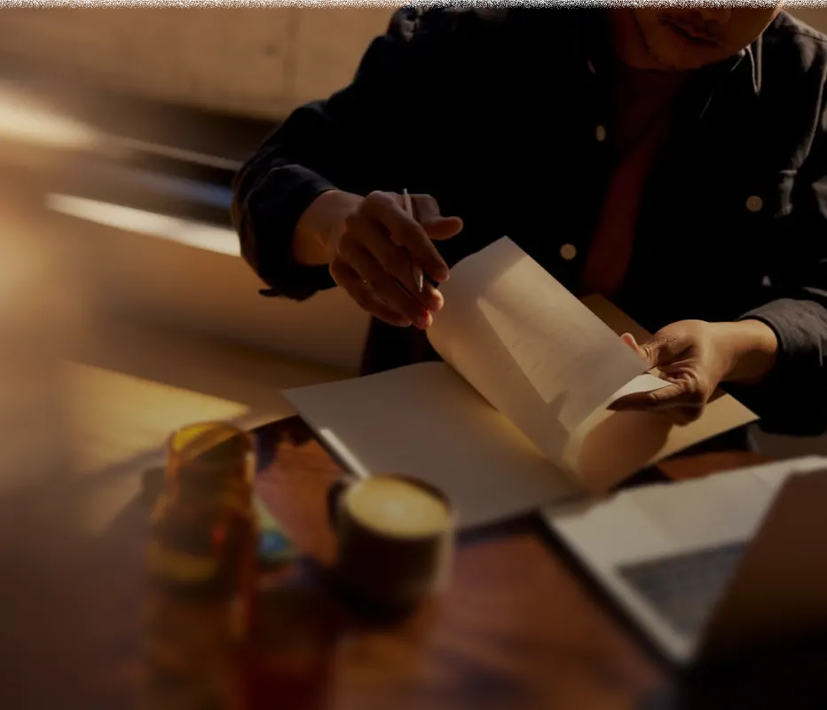 Person flipping through pages of a notebook or book at a wooden table with a pen in hand, a cup of coffee, and a laptop nearby in warm lighting.