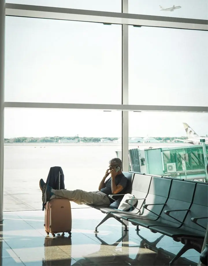 Homem ao telefone no saguão do aeroporto, com a mala ao lado, enquanto um avião decola